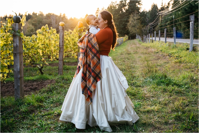 mother wearing wedding dress nuzzles baby while standing in field at sunset