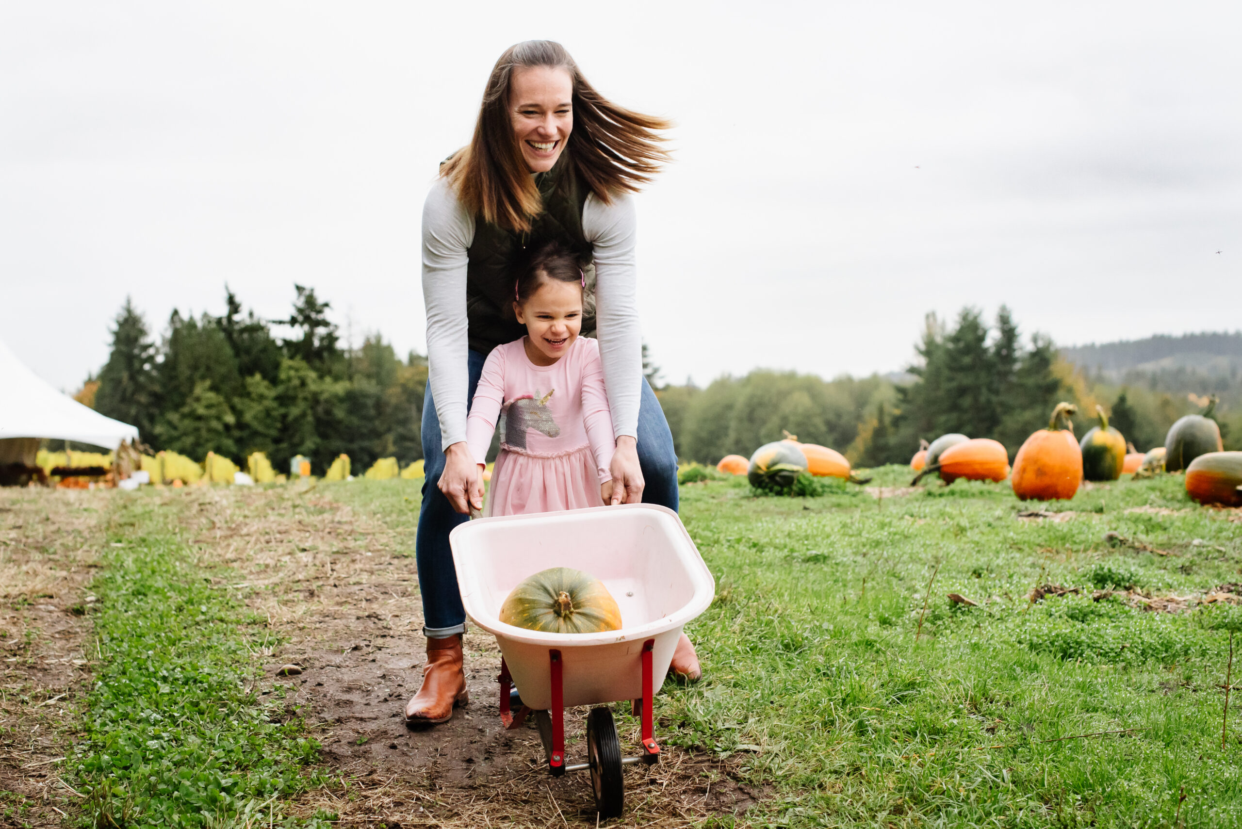 Mom and daughter laughing while pushing a wheelbarrow at a Kitsap County pumpkin patch, lifestyle family photography near Silverdale and Poulsbo