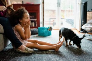 Young girl yawning on the living room floor as the family dog sniffs her toes, candid in-home documentary family photography in Poulsbo