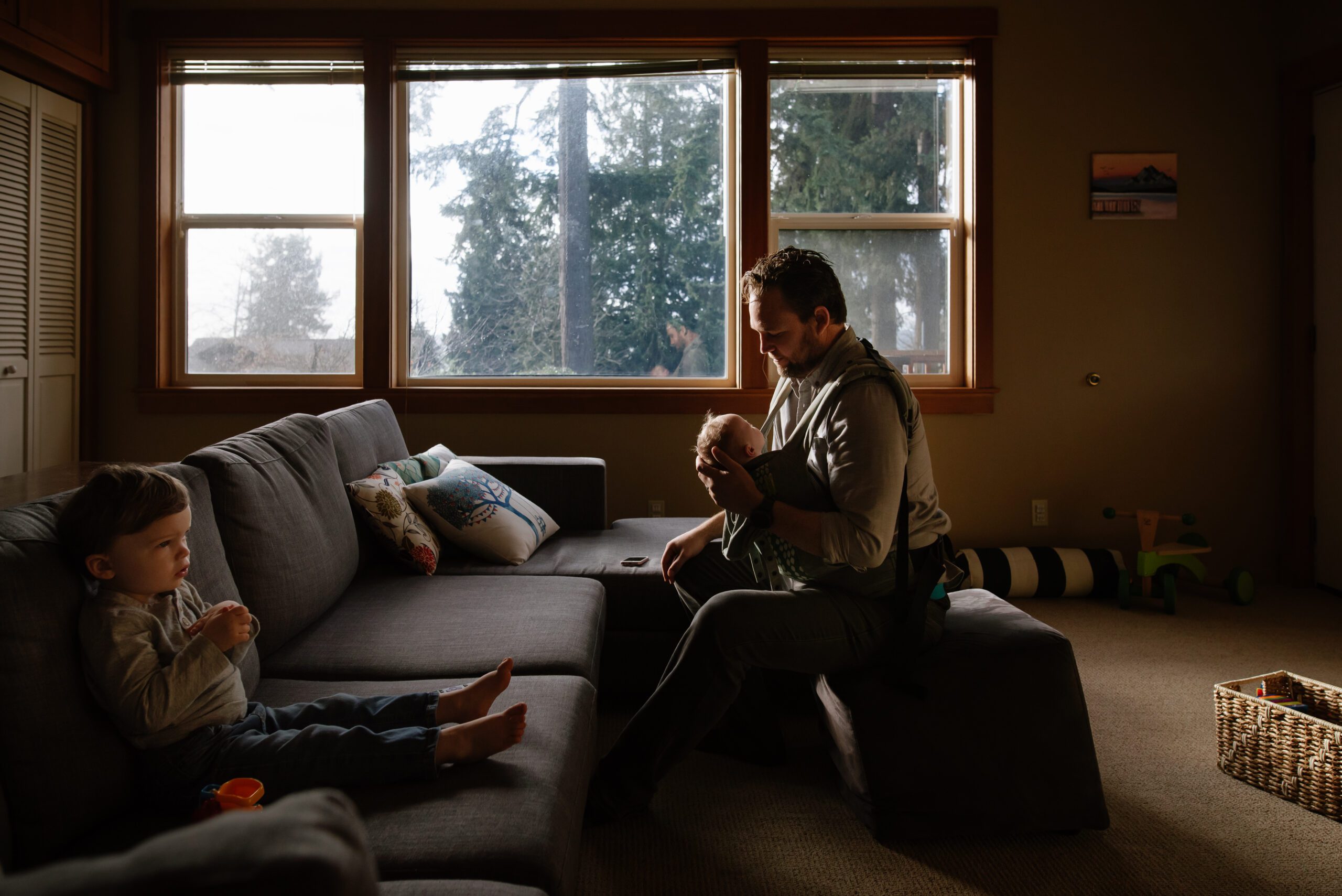 Dad holding a newborn in window light while a toddler looks on from the couch, in-home documentary family photography on Bainbridge Island