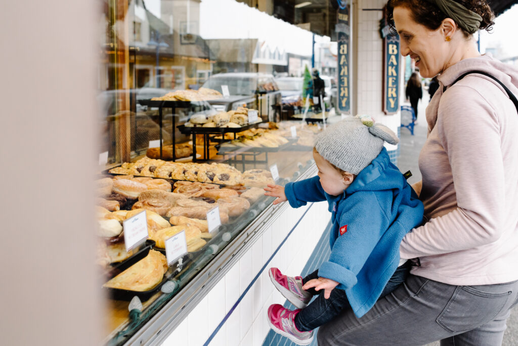Mom holding toddler up to a bakery display window in downtown Poulsbo, documentary lifestyle family photography in Kitsap County